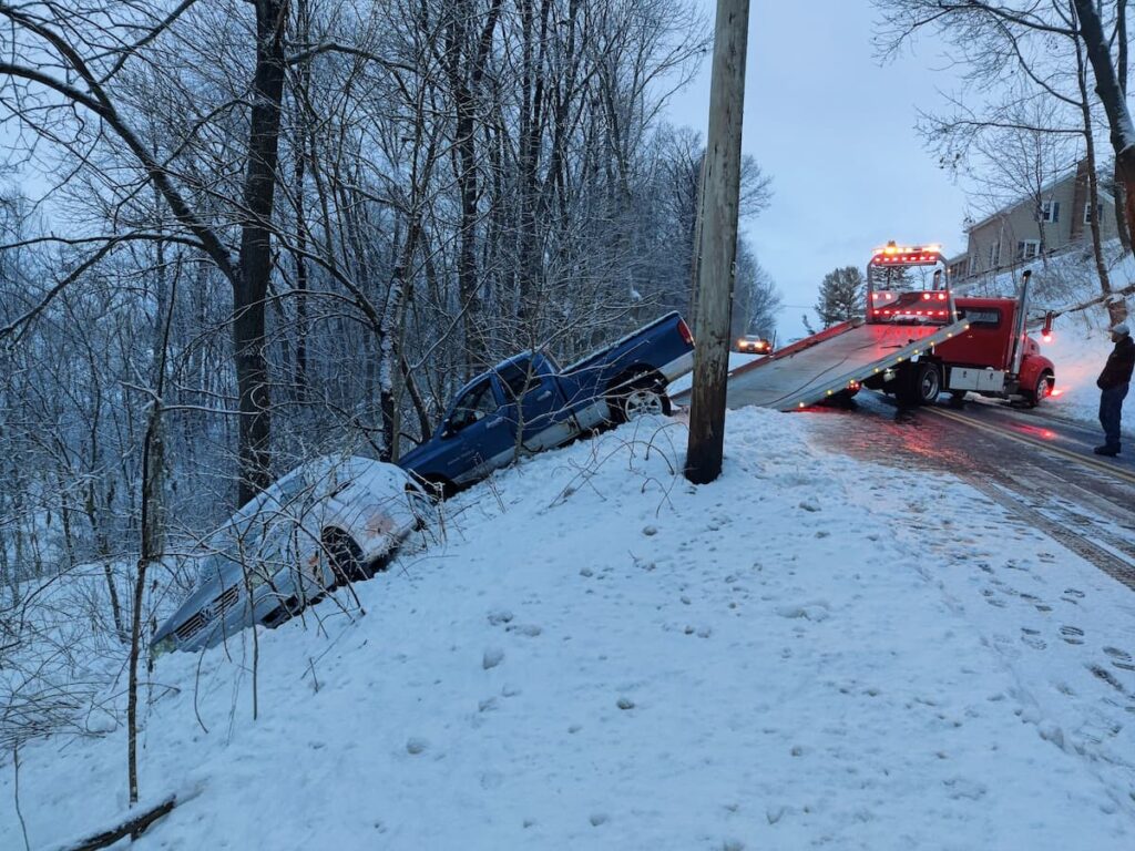 Tow truck preparing to recover a pickup and car off the road in snowy conditions.